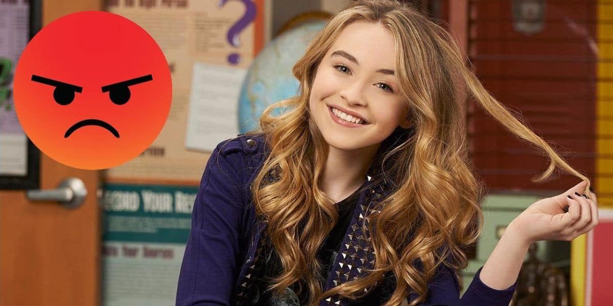 A person with long, wavy hair smiles while holding a strand of hair, resembling Sabrina Carpenter amidst recent controversy. They wear a dark jacket over a patterned shirt. An angry face emoji is on the left. The background features posters and a partially visible door.