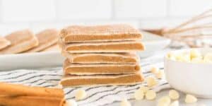 A stack of cinnamon-laced cookies sprinkled with sugar on a white plate, beside a bowl of white chocolate chips, set against a kitchen tile backdrop.