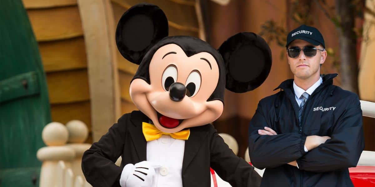 A person in a Mickey Mouse costume poses for the camera with an arm outstretched, while a security guard wearing sunglasses and a cap labeled "Security" stands beside them with arms crossed. The background features vibrant, whimsical buildings.