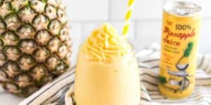 A glass of creamy pineapple smoothie topped with swirls, accompanied by a straw, standing next to a fresh pineapple and a can of pineapple juice on a striped cloth.