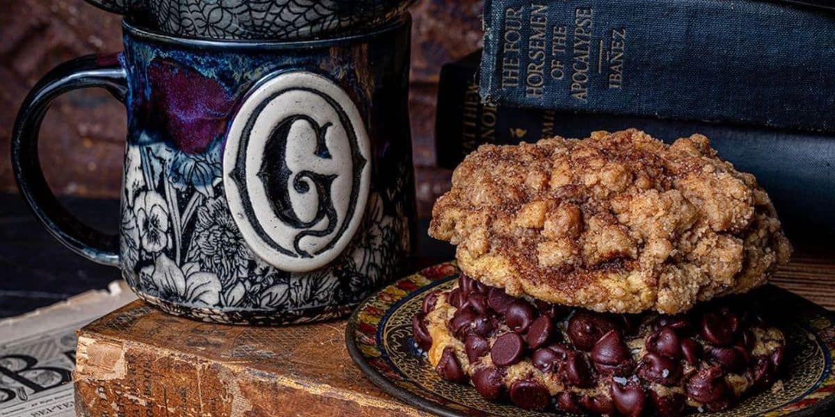 A close-up of a dessert and a mug. The dessert consists of a large chocolate chip cookie topped with a crumbly cinnamon streusel. The decorative mug has a letter "G" on it. In the background, there are several hardcover books stacked.