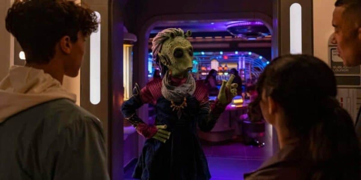 A green alien character is gesturing with one hand in a sci-fi themed room, speaking to three Star Wars fans standing in a doorway. The room behind is illuminated with colorful neon lights, creating a futuristic ambiance.