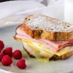 A Monte Cristo breakfast sandwich dusted with powdered sugar, served with raspberry jam, fresh raspberries, and a glass of milk on a light-colored table.