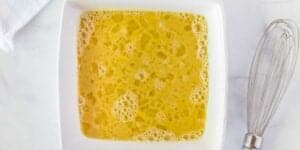 A square white bowl containing a beaten egg mixture beside a metal whisk, placed on a white surface. The focus is on the vibrant, frothy yellow of the eggs, essential for a Monte Cristo Breakfast Sandwich Recipe.