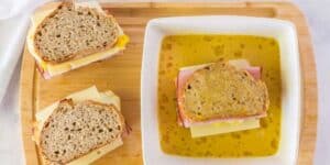 A bowl of yellow soup with a Monte Cristo sandwich half submerged, alongside a cutting board with two more Monte Cristo sandwich halves stacked with ham and cheese, all on a marble surface.