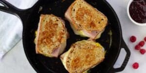 Three Monte Cristo sandwiches with ham in a cast iron skillet, next to a dish of raspberry jam and fresh raspberries on a marble countertop.