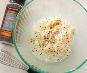 A glass bowl containing a mixture of spices and liquid ingredients for a Ronto Wrap recipe, with a whisk and a container of black pepper on the side.