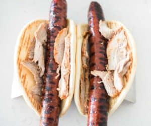 Two open sandwich-style pita breads with grilled sausages and slices of roast pork, inspired by the Ronto Wrap recipe, on a white marble surface.