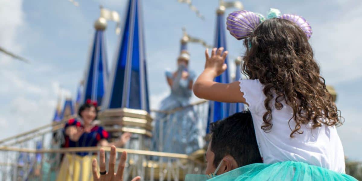 A child wearing Minnie Mouse ears sits on an adult's shoulders, waving at costumed characters on a blue and white float in an amusement park parade. Blue sky and clouds are visible in the background.