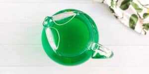 A glass pitcher filled with green liquid stands on a white wooden surface, viewed from above. Part of a white and green patterned cloth is visible in the top right corner.