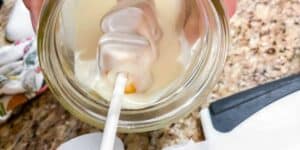 A marshmallow on a stick, resembling Tigger Tails, is being dipped into a creamy white coating inside a glass jar. The scene is set on a speckled granite countertop with a knife and a floral cloth nearby.