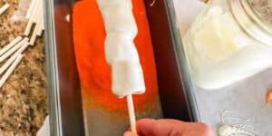 A hand is dipping a Tigger Tail—a white chocolate-covered food item on a stick—into orange-colored sugar or sprinkles inside a rectangular pan. Surrounding are a jar of white chocolate and additional white-covered items on sticks.