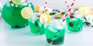 Three glasses of bright green beverage served with ice cubes, lemon slices on the rim, red cherries, and striped red and white paper straws. In the background, there's a large pitcher filled with the same green drink, ice, and a lemon slice on the rim.