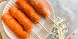 Four crispy, breaded corn dogs on sticks are placed on a white paper plate. Beside the plate, reminiscent of Tigger Tails, there is a spill of white shredded cheese and some crumbs on parchment paper.