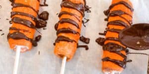 Three orange ice pops on sticks, resembling Tigger Tails, are drizzled with chocolate sauce. The ice pops are laid out on parchment paper, and a spoon is in the process of adding more chocolate to the treat on the right.