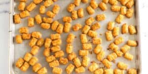 A baking sheet filled with evenly spaced golden-brown tater tots, fresh out of the oven, ready to become totchos. The crispy tots are slightly glistening, arranged randomly across the tray, showcasing their irresistible texture. The background is a marble countertop.