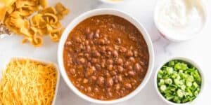 A bowl of chili with beans sits in the center, surrounded by small dishes of shredded cheese, corn chips, chopped green onions, sour cream, and grated cheese on a white surface—perfect for creating your own delicious totchos.