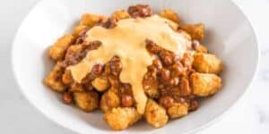 A white bowl filled with crispy tater tots, transformed into delectable totchos, topped with warm chili and melted cheese. The dish appears appetizing with the contrasting textures of crunchy tots and the creamy chili-cheese mix.