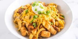A white bowl filled with Frito pie, featuring corn chips, chili, beans, shredded cheddar cheese, a dollop of sour cream, and garnished with chopped green onions. Inspired by Totchos, this dish is presented against a plain, light-colored background.