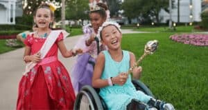 Three girls in princess costumes, one in a wheelchair, joyfully playing outdoors at sunset, with lush greenery in the background.