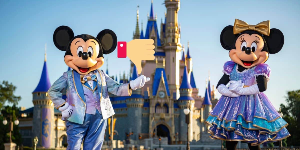 Mickey Mouse and Minnie Mouse, wearing formal outfits, stand in front of Cinderella's Castle at Disneyland. Mickey is pointing to a large thumbs-down emoji floating between them while Minnie holds a proud pose.