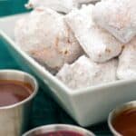 A plate of powdered sugar-coated beignets accompanied by three small dipping sauces in stainless steel cups, set on a green textured background.