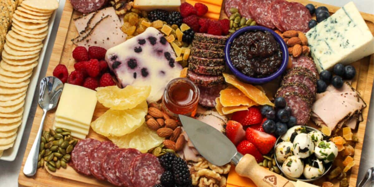 A wooden charcuterie board filled with various types of cheese, meats, fruits, nuts, and spreads. Sliced salami, berries, dried fruits, almonds, and crackers are present, along with cheese cubes and a bowl of jam. A cheese knife and spreader lie on the board.