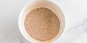 A close-up view of a white bowl filled with fine, light brown powder, placed on a marble surface. the texture of the powder appears smooth and uniform.