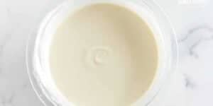 A clear glass bowl containing a smooth, creamy beige sauce or dressing, viewed from above on a marble countertop.