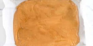 A uniform layer of smooth peanut butter fudge spread out on a parchment paper-lined surface. the fudge has a creamy, light brown color and a glossy texture.