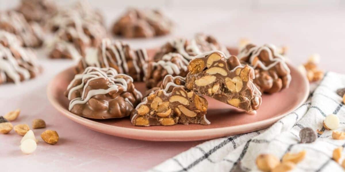 Close-up of a pink plate filled with clusters of chocolate-covered peanuts drizzled with white chocolate. One cluster is cut in half, revealing the peanut center. Some loose peanuts and chocolate chips are scattered around the plate on a pink surface.