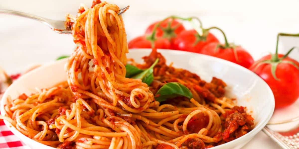 A bowl of spaghetti topped with a rich tomato sauce and garnished with fresh basil leaves. A fork is lifting a portion of the pasta. In the background, there are ripe red tomatoes on the vine. The scene is bright and appetizing.