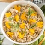A bowl of rice mixed with ground meat, topped with bright yellow flower petals and garnished with sprigs of rosemary. The bowl is placed on a green checkered cloth with a gold spoon beside it, and additional rosemary sprigs are on the table.