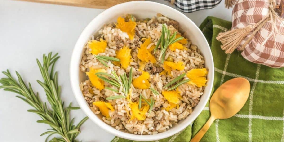 A bowl of rice mixed with ground meat, topped with bright yellow flower petals and garnished with sprigs of rosemary. The bowl is placed on a green checkered cloth with a gold spoon beside it, and additional rosemary sprigs are on the table.