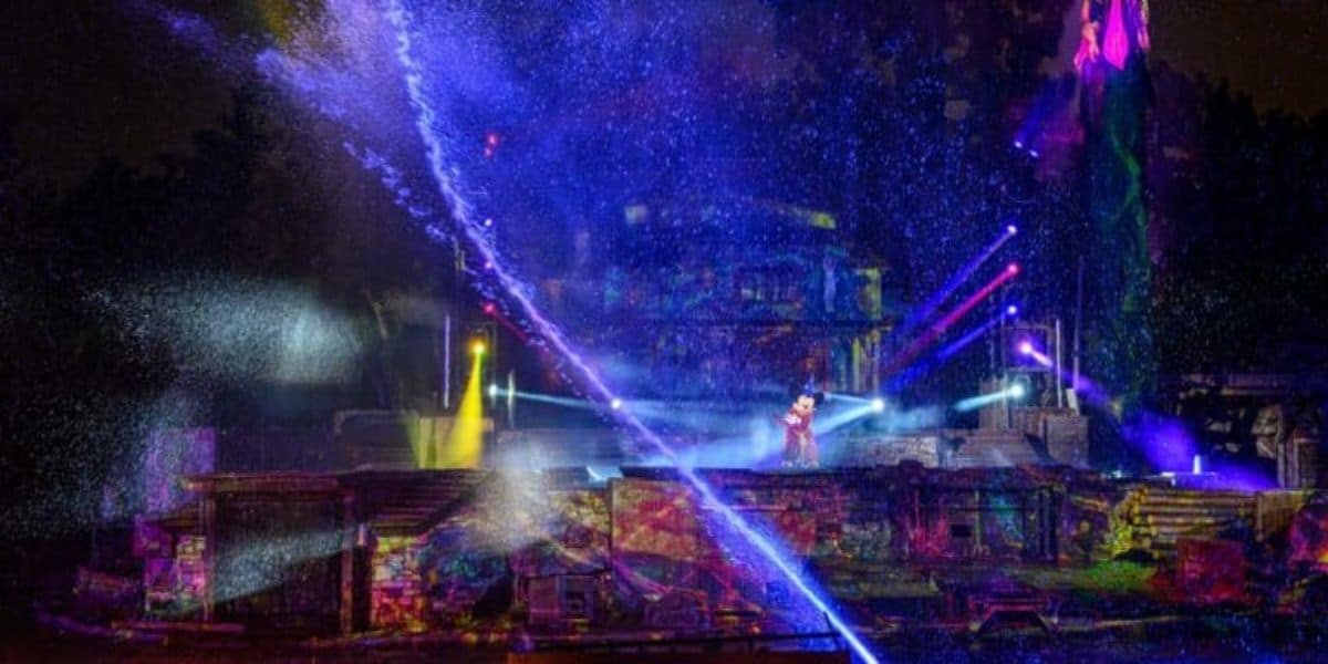 A vividly lit outdoor stage show at night featuring colorful lights, mist, and water sprays. The backdrop includes multicolored lighting effects creating a magical, dreamlike atmosphere. A character, possibly a performer, stands illuminated in the center.