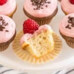 A white cake stand displays several vanilla cupcakes topped with pink frosting. Some cupcakes are garnished with chocolate sprinkles, while others are topped with a ripe raspberry. One cupcake is partially eaten, showing its soft and fluffy interior.