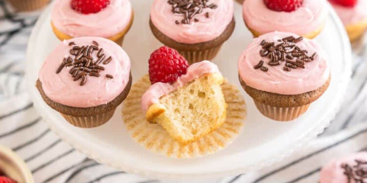 A white cake stand displays several vanilla cupcakes topped with pink frosting. Some cupcakes are garnished with chocolate sprinkles, while others are topped with a ripe raspberry. One cupcake is partially eaten, showing its soft and fluffy interior.