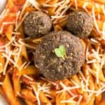 A bowl of penne pasta coated in red sauce topped with shredded cheese. There are three meatballs arranged on top of the pasta, with one large meatball in the center and two smaller ones positioned to resemble a face. A small green leaf adorns the large meatball.