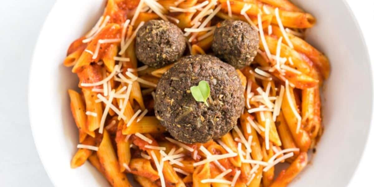 A bowl of penne pasta coated in red sauce topped with shredded cheese. There are three meatballs arranged on top of the pasta, with one large meatball in the center and two smaller ones positioned to resemble a face. A small green leaf adorns the large meatball.
