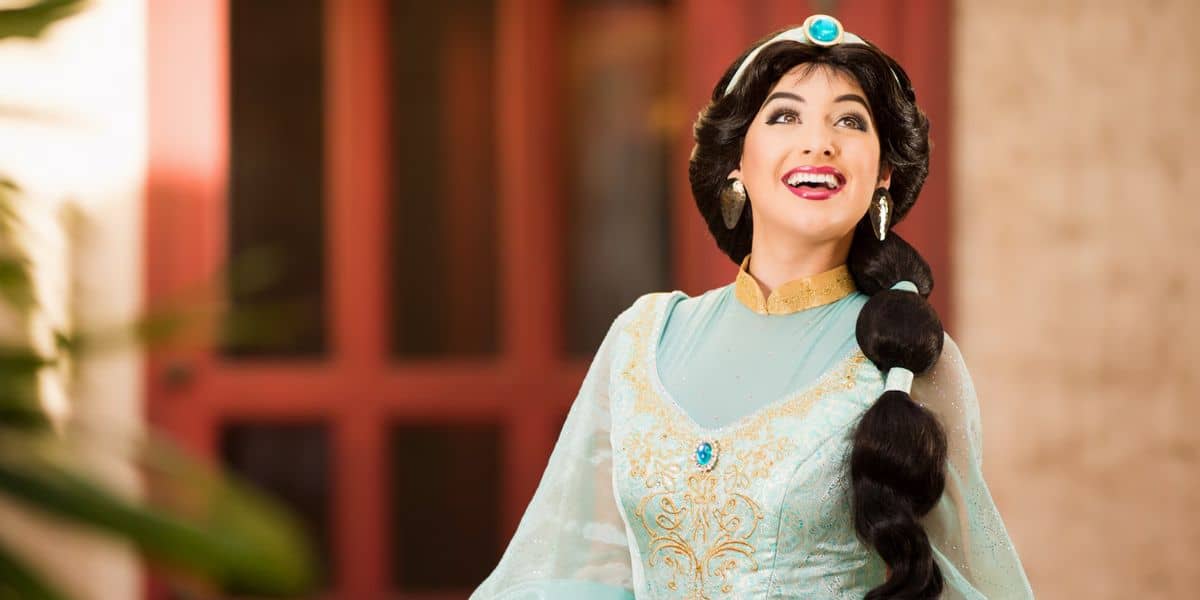 A woman dressed in a light blue and gold costume with a jeweled headband smiles and looks upwards. She has long, dark, braided hair and wears golden jewelry. The background is softly blurred with hints of a red door and greenery.