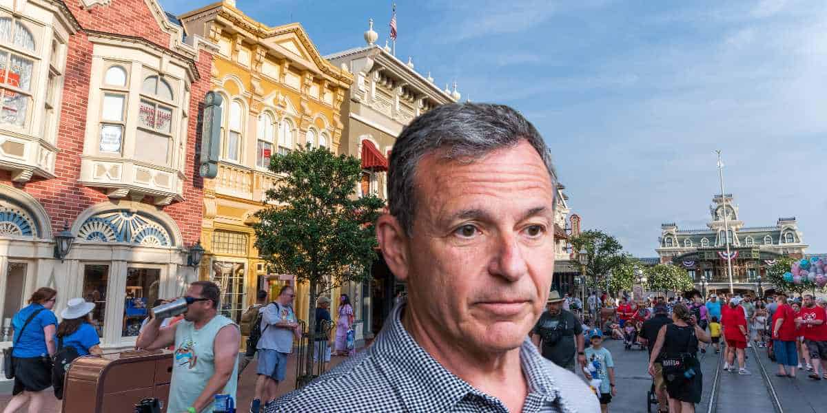 Bob Iger with a serious expression standing in a crowded Disney theme park with classic style buildings in the background.