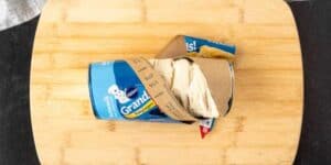A Pillsbury biscuit dough container, partially opened with the dough expanding out, prepped on a wooden cutting board for a fried biscuits recipe.
