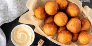 A basket of golden brown fried biscuit holes served with a side of honey for dipping, presented on a patterned cloth background.