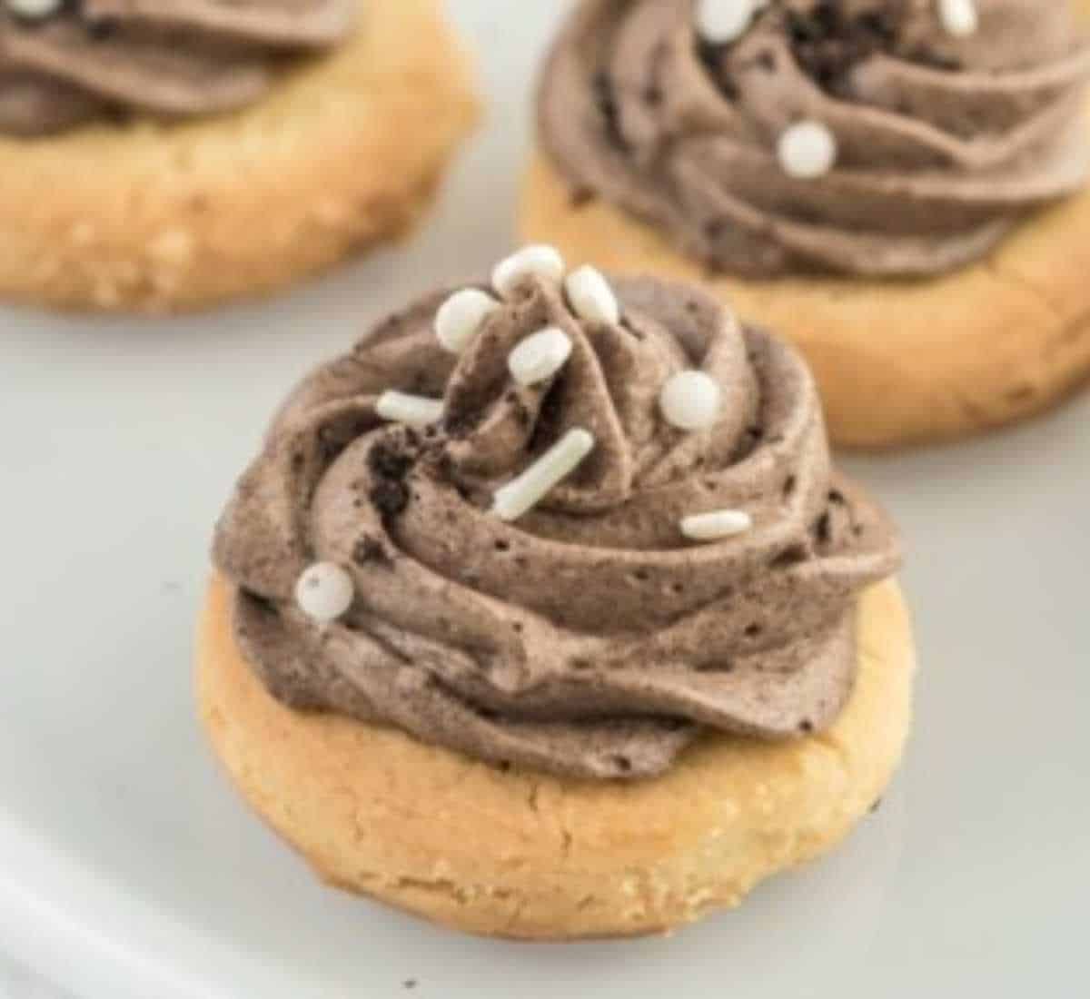 Three cookies topped with a swirl of grey stuff and sprinkled with white beads, displayed on a light gray plate.