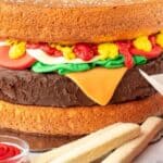 A creative cake designed to look like a hamburger, with layers resembling buns, a patty, lettuce, tomato, and cheese. It is accompanied by finger-shaped cookies arranged like fries, with a small cup of red icing simulating ketchup on the side.