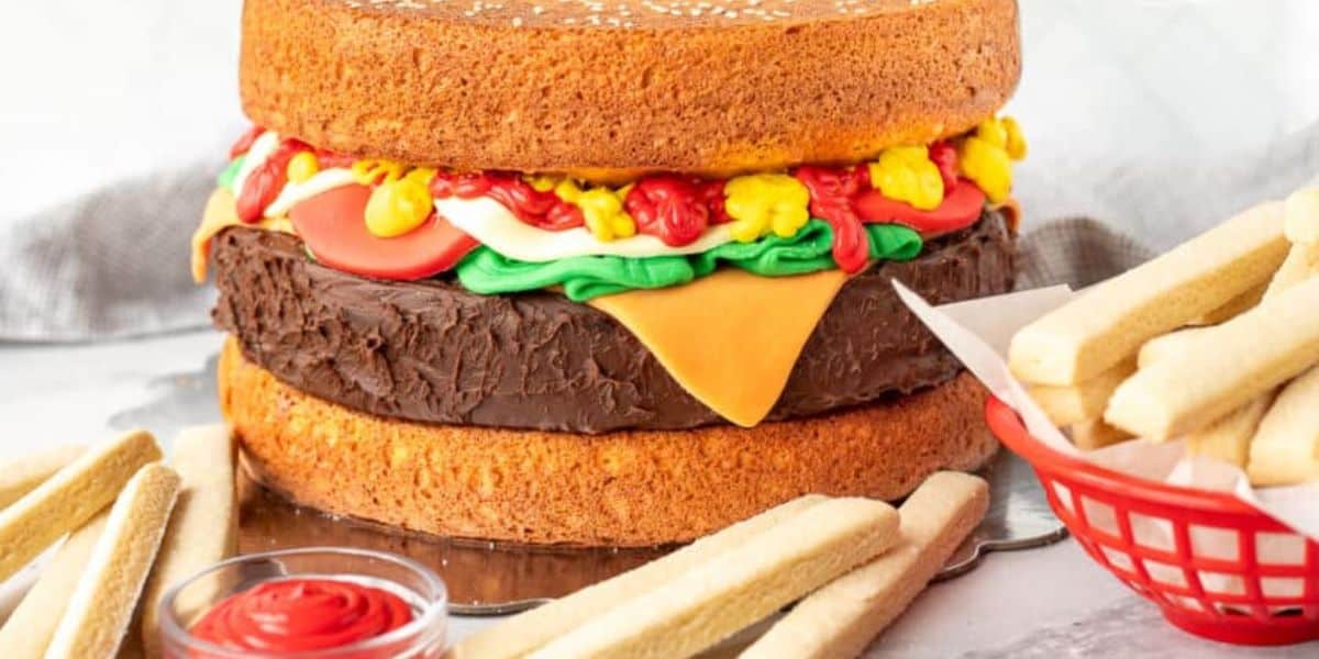 A creative cake designed to look like a hamburger, with layers resembling buns, a patty, lettuce, tomato, and cheese. It is accompanied by finger-shaped cookies arranged like fries, with a small cup of red icing simulating ketchup on the side.