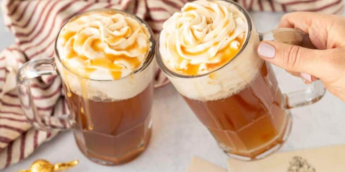 Two glass mugs filled with a frothy, caramel-topped beverage, likely butterbeer, are being clinked together. The drinks have whipped cream and caramel drizzle on top. A red and white striped cloth and a golden spoon are in the background.