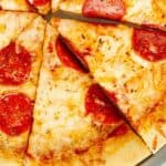 A close-up view of a pepperoni pizza with a golden crust and melted cheese, topped with evenly spaced slices of pepperoni. The pizza is pre-sliced into eight pieces and served on a round metal pan.