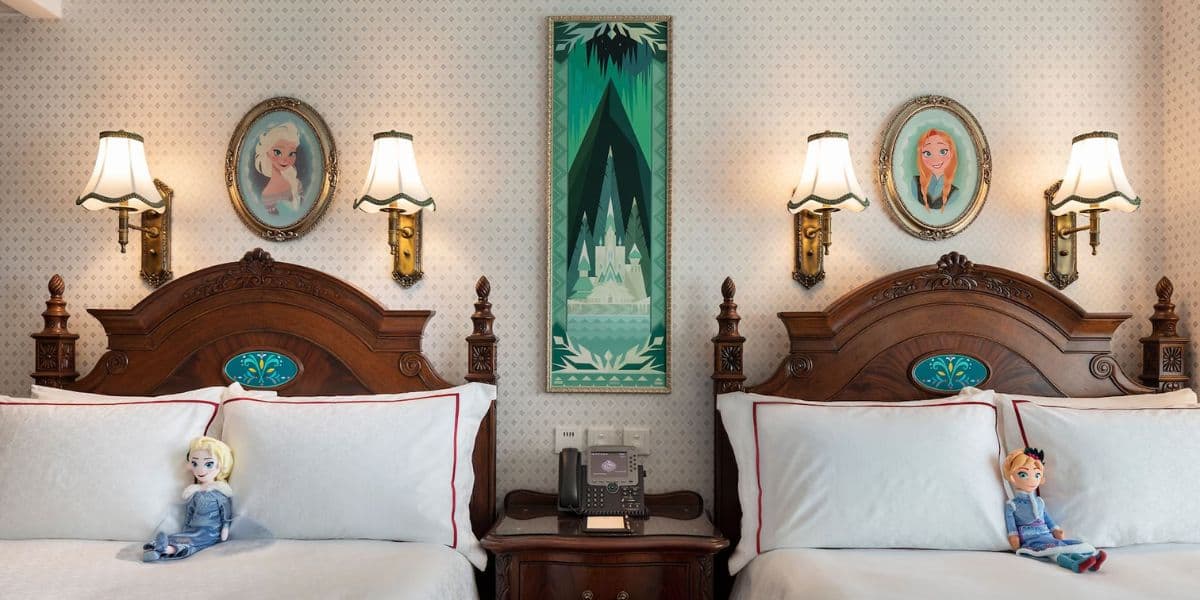 A cozy hotel room features two wooden beds with white and blue linens, each adorned with an Elsa and Anna doll from Frozen. Between the beds is a phone on a nightstand. Above the beds are paintings of Elsa and Anna, with a central framed artwork of a castle.
