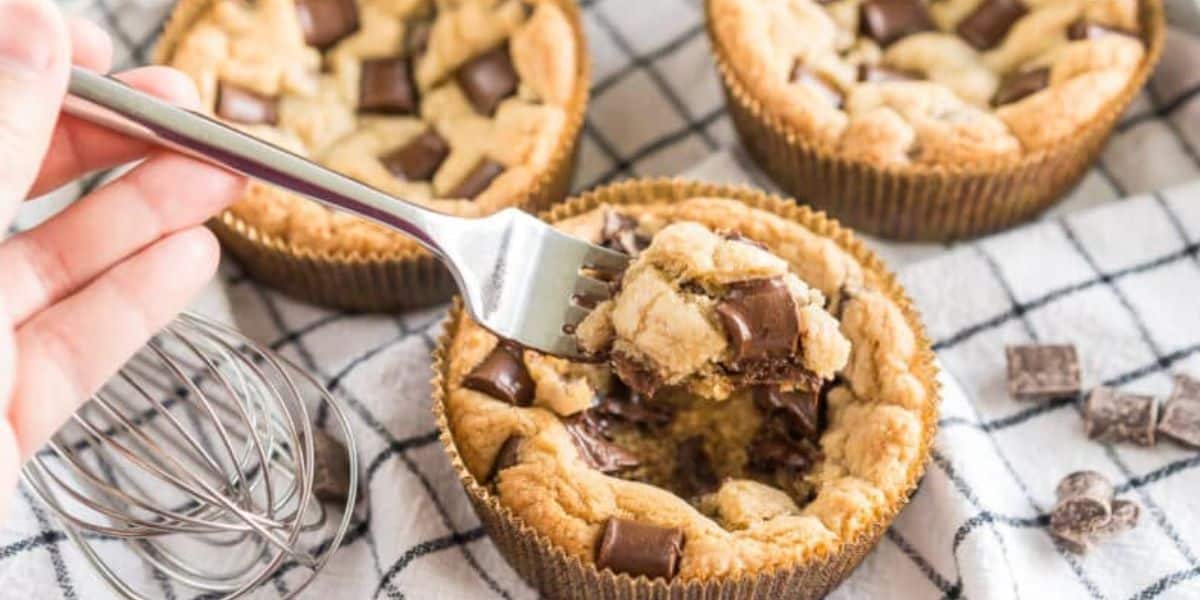 A hand holding a fork about to take a bite from a large cookie cup filled with melted chocolate chunks. Two similar cookie cups and a small whisk are on a checkered cloth, with extra chocolate chunks scattered nearby.